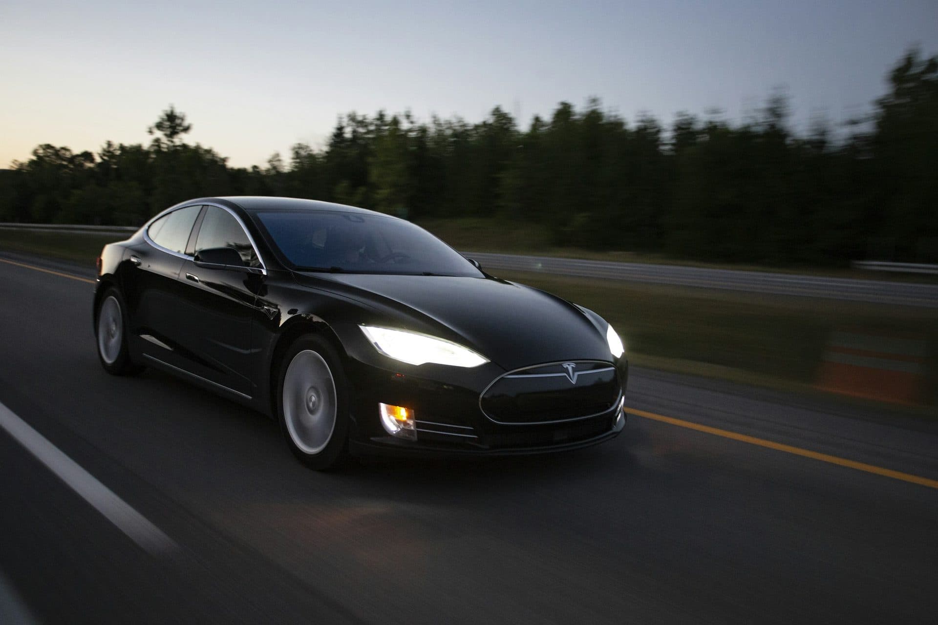 Black Tesla Model S driving on a highway at dusk with its headlights on