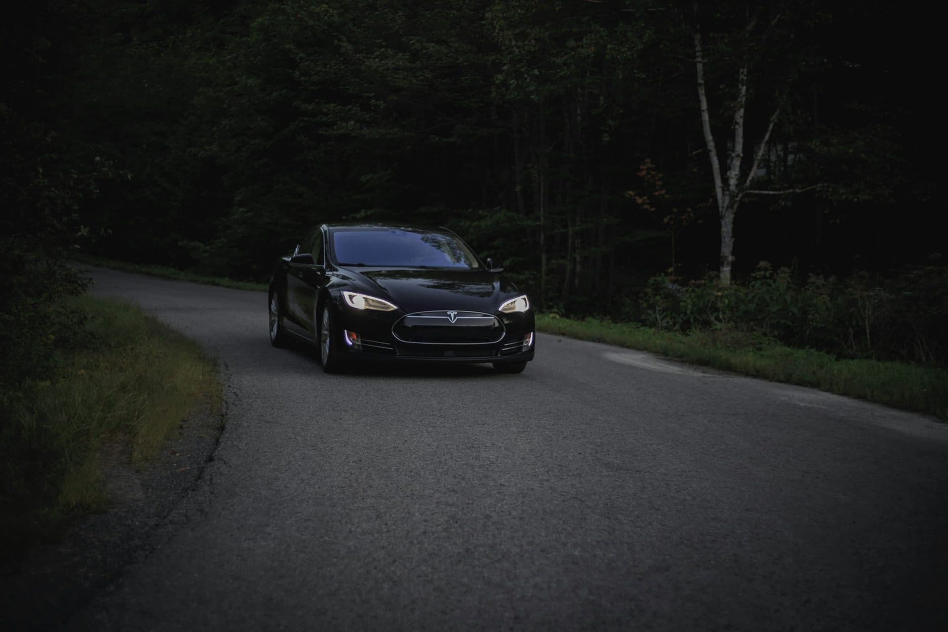 Black Tesla Model S with headlights on, on a dark tree-lined road in the evening